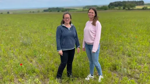 Anna Biesty Anna Biesty, dressed in a pink jumper and jeans, stands in a field, planted with wildflower strips, with her sister. There are trees on the horizon.