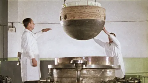 Getty Images The image shows two individuals in white lab coats lowering the descent capsule of the Soviet space probe Venera 5 or 6 into a large, cylindrical metal structure to test the heat resistance. The capsule is spherical with a dark bottom half and a lighter top half, featuring several circular holes. 
