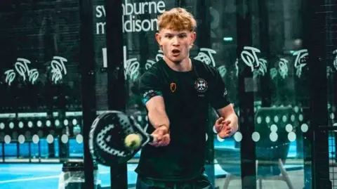Daisy Whitlie A young man in sports kit strikes a ball with a padel racquet. He has an expression of intense concentration and looks to be exerting himself as the forehand swing connects.