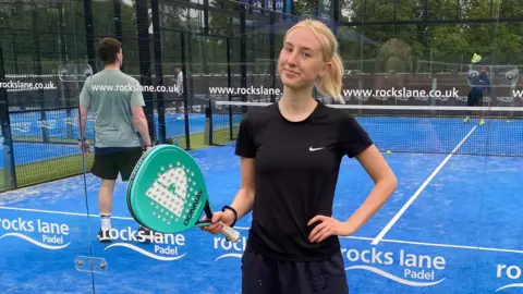 Rosie Hepworth Rosie is standing in front of a blue astro padel court. She is wearing a black t-shirt and matching tracksuit bottoms and we can only see her from the waist up. She is holding a turquoise padel racket in one hand and the other hand is on her hip. Rosie has shot blonde hair, which has been tied back into a low ponytail. She is smiling into the camera with no teeth. Behind her are two men playing on the court, who can be seen through the glass walls. There is white branding written on the glass.