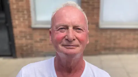 Mousumi Bakshi/BBC Paul Davis is looking at the camera with a light smile. He is wearing a white T-shirt and has short grey hair. He is standing in front of a red bricked building. 