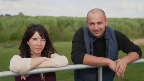 Gwyndaf Hughes/BBC A man wearing a black t-shirt and blue gilet stands next to a woman wearing a white t-shirt and red gilet. They are leaning over a metal fence in a green field, with their arms resting on the fence. 