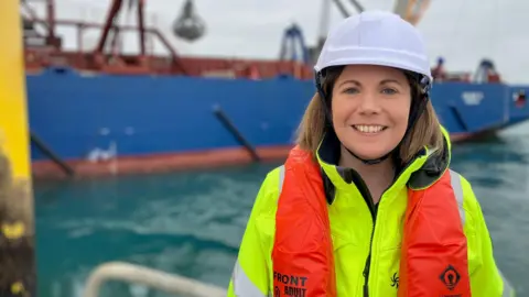 A woman is smiling at the camera - she has blonde hair and is wearing a hi-vis yellow jacket, an orange life vest and a white helmet. Behind her is a blue boat and the sea