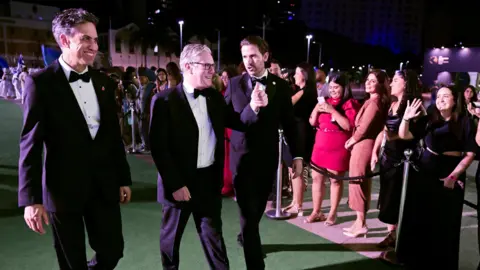 Reuters Ed Miliband and Sir Keir Starmer walk along a green carpet with a man in a tuxedo. Starmer, in the middle, gives a waving member of the nearby crowd a thumbs-up.