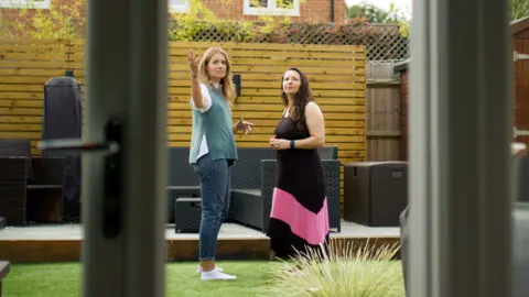Two women standing and talking in a garden area, viewed through a glass door. The garden has a wooden deck with dark outdoor furniture, a wooden fence with lattice on top, and green grass in the foreground. There is a plant with spiky leaves near the bottom of the image, and a brick building partially visible in the background.