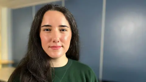 Stephanie Metzger wearing a dark green sweatshirt with long, dark hair, standing indoors in front of a blue wall with vertical panel lines. The background includes a blurred light-colored surface on the left side, suggesting a table or counter.
