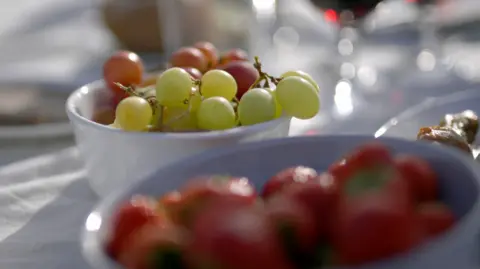Close-up of a white bowl filled with green and red grapes on a table covered with a white cloth. In the foreground, another bowl contains red strawberries, slightly out of focus. The background shows blurred glassware and other picnic items, suggesting an outdoor dining setting in bright natural light.
