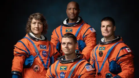 NASA The image shows four astronauts in bright orange space suits posing for a formal group portrait against a dark background. The suits are bulky with blue joints, straps and pockets, emphasising their technical design. Three astronauts stand behind while one is seated or crouching in front, all facing the camera. Their faces are blurred into smooth ovals so no expressions can be seen, keeping the focus on the uniforms. Mission-style patches and flags, including US and Canadian flags, are visible on their arms and chests. Soft, focused lighting makes the vivid orange suits stand out dramatically.